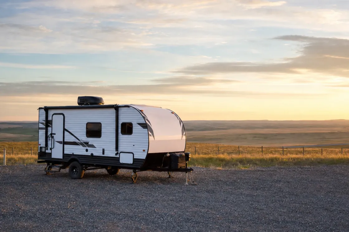 A lightweight camping trailer parked on a gravel dealership pad at golden hour with open Alberta prairie and soft evening skies