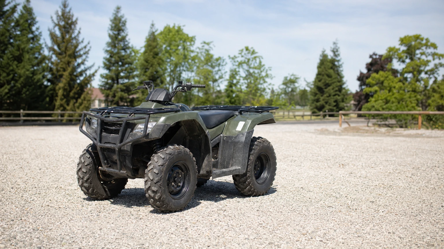 A clean used four-wheel ATV parked alone on a gravel lot with trees and open sky behind it
