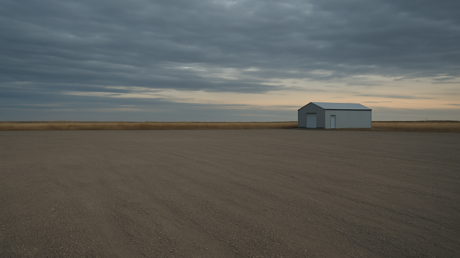 A wide overcast Alberta sky over an empty flat gravel dealership lot with a plain metal building small on the horizon